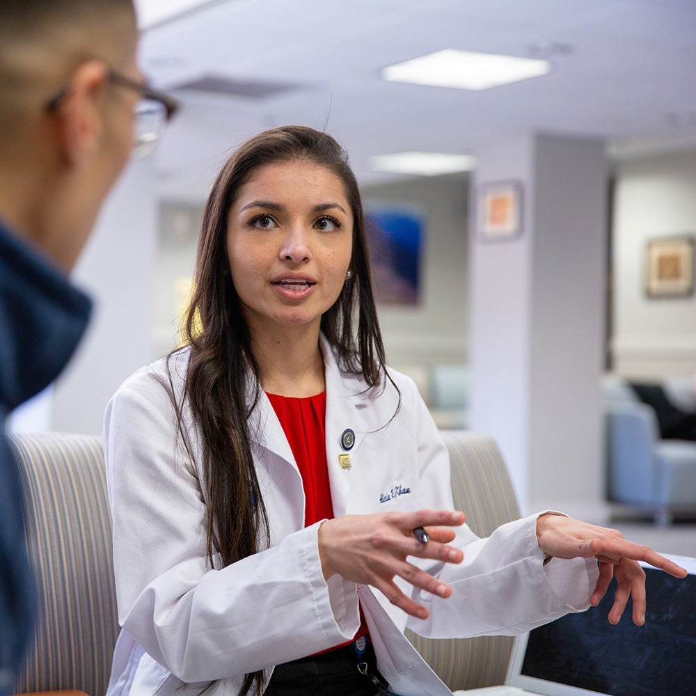 A medical student in a white coat speaks to another med student