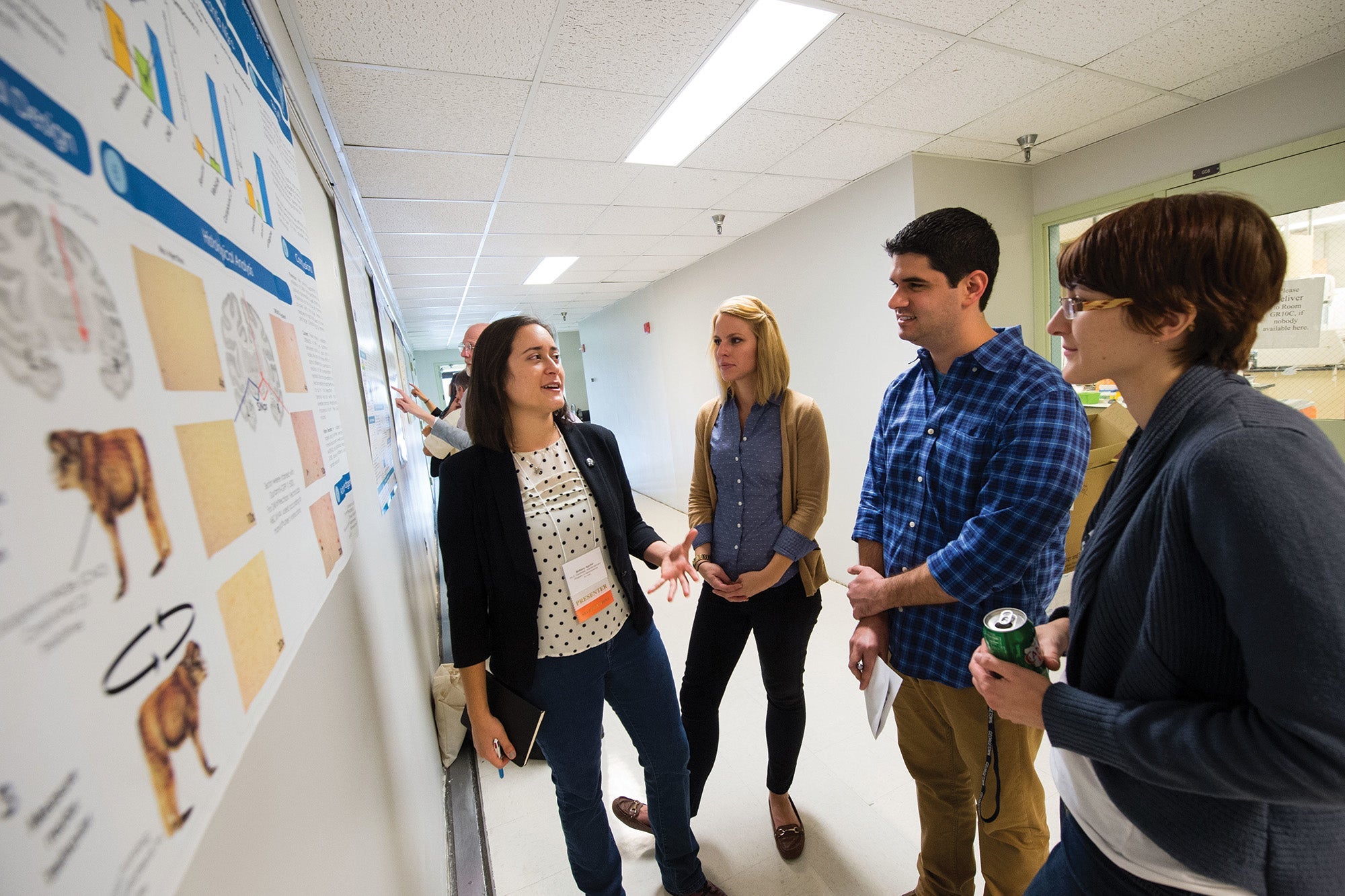 four students stand in a hallway as one explains her research poster on the wall