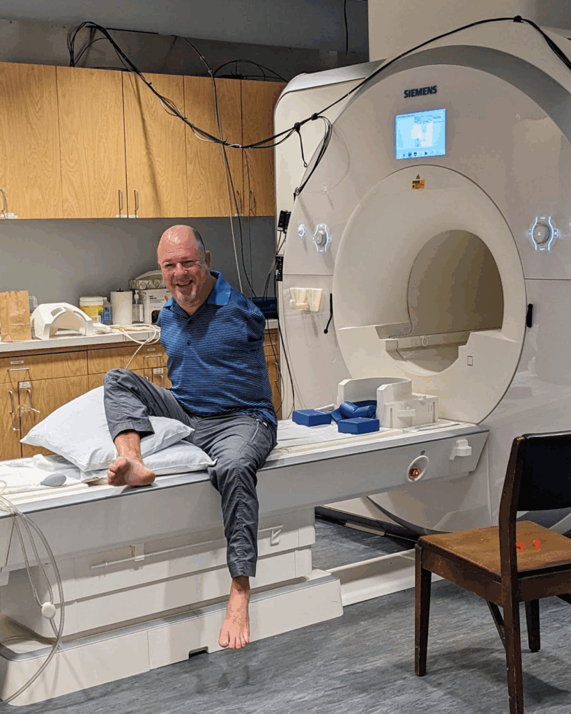 Alvin Law has no arms and bare feet, one of which is flexed and propped on pillows. He sits upright on the bed of an MRI machine.