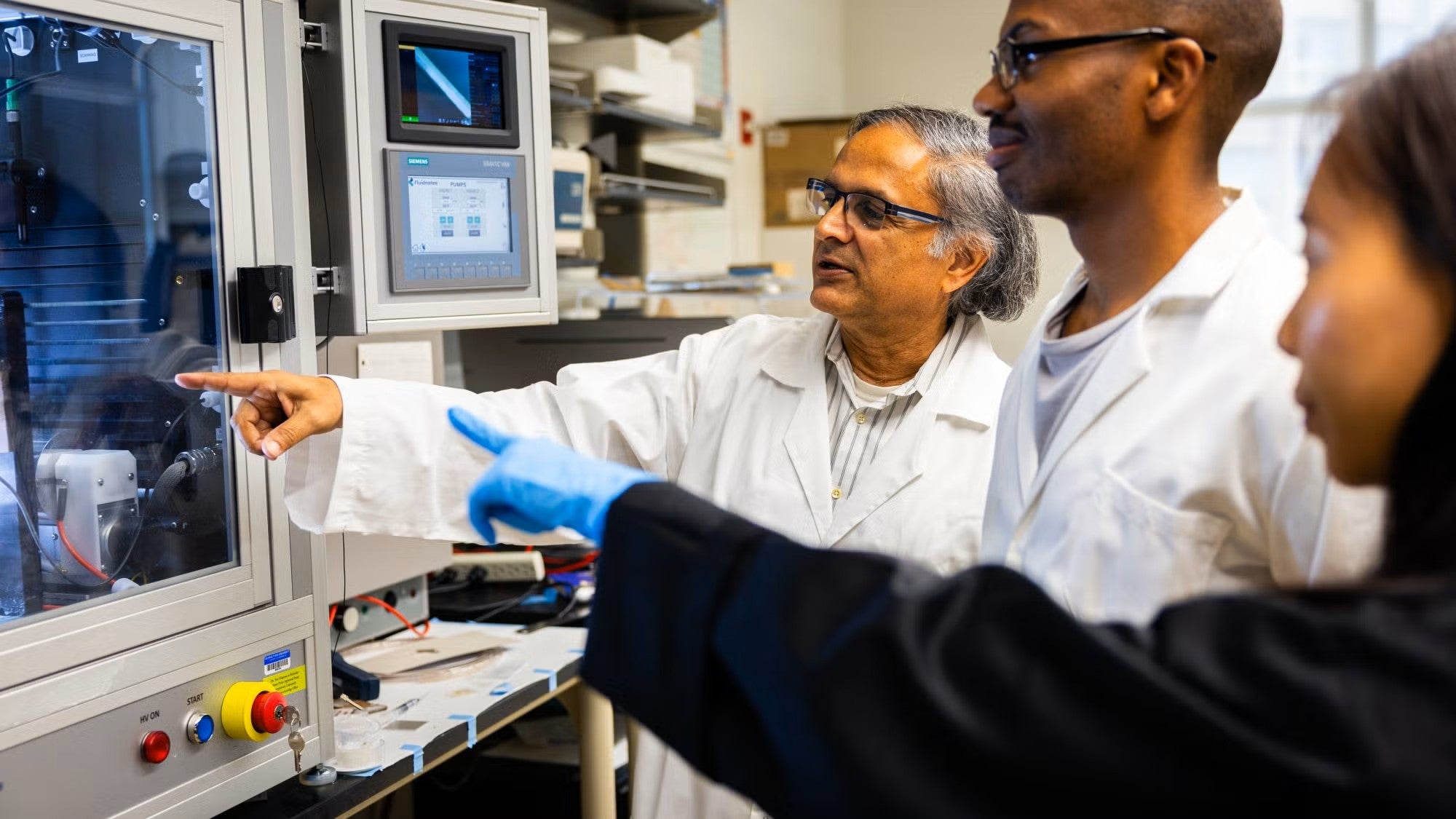 Three researchers point to an object under a vent hood in the lab