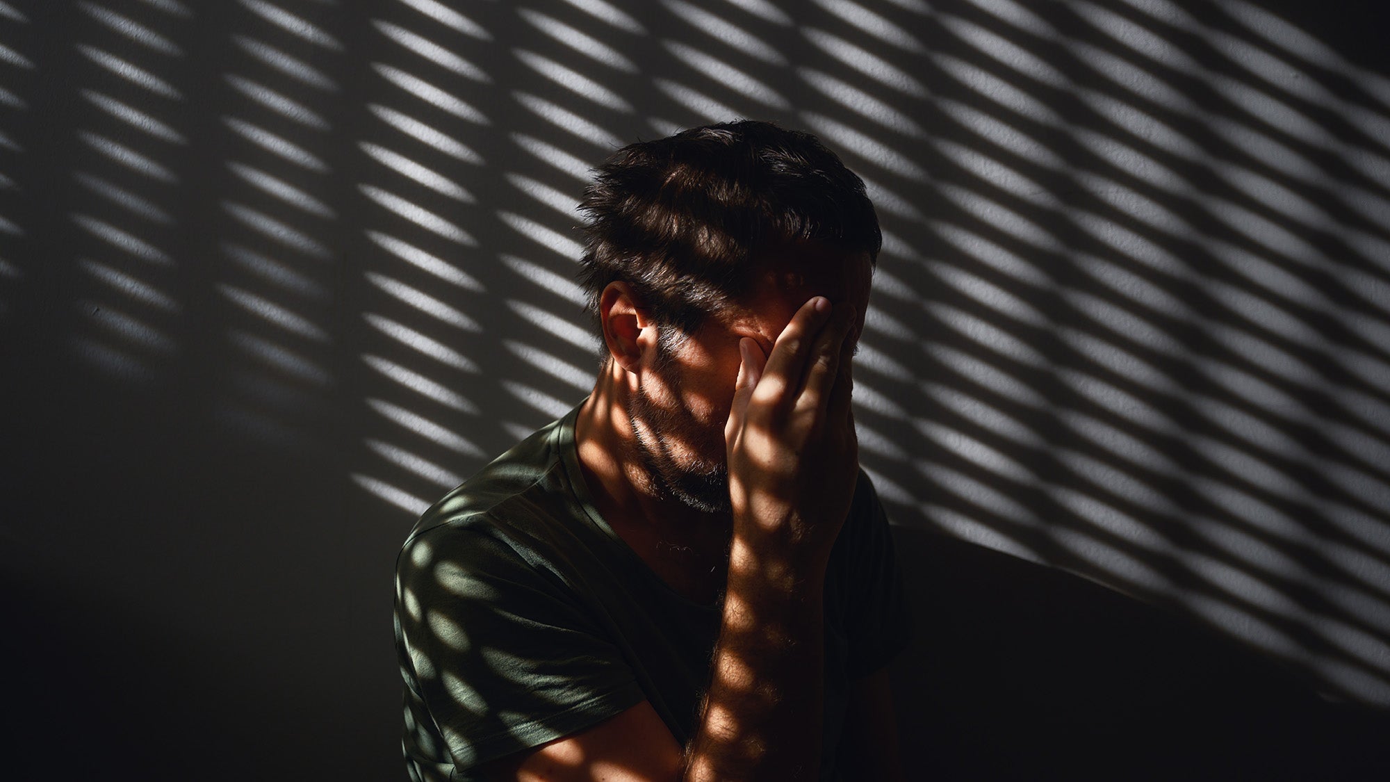 A man sits in a darkened room, holding his head in pain