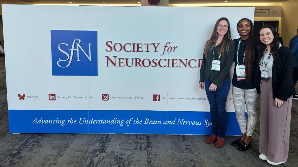 Three individuals stand in front oa sign for the Society for Neuroscience