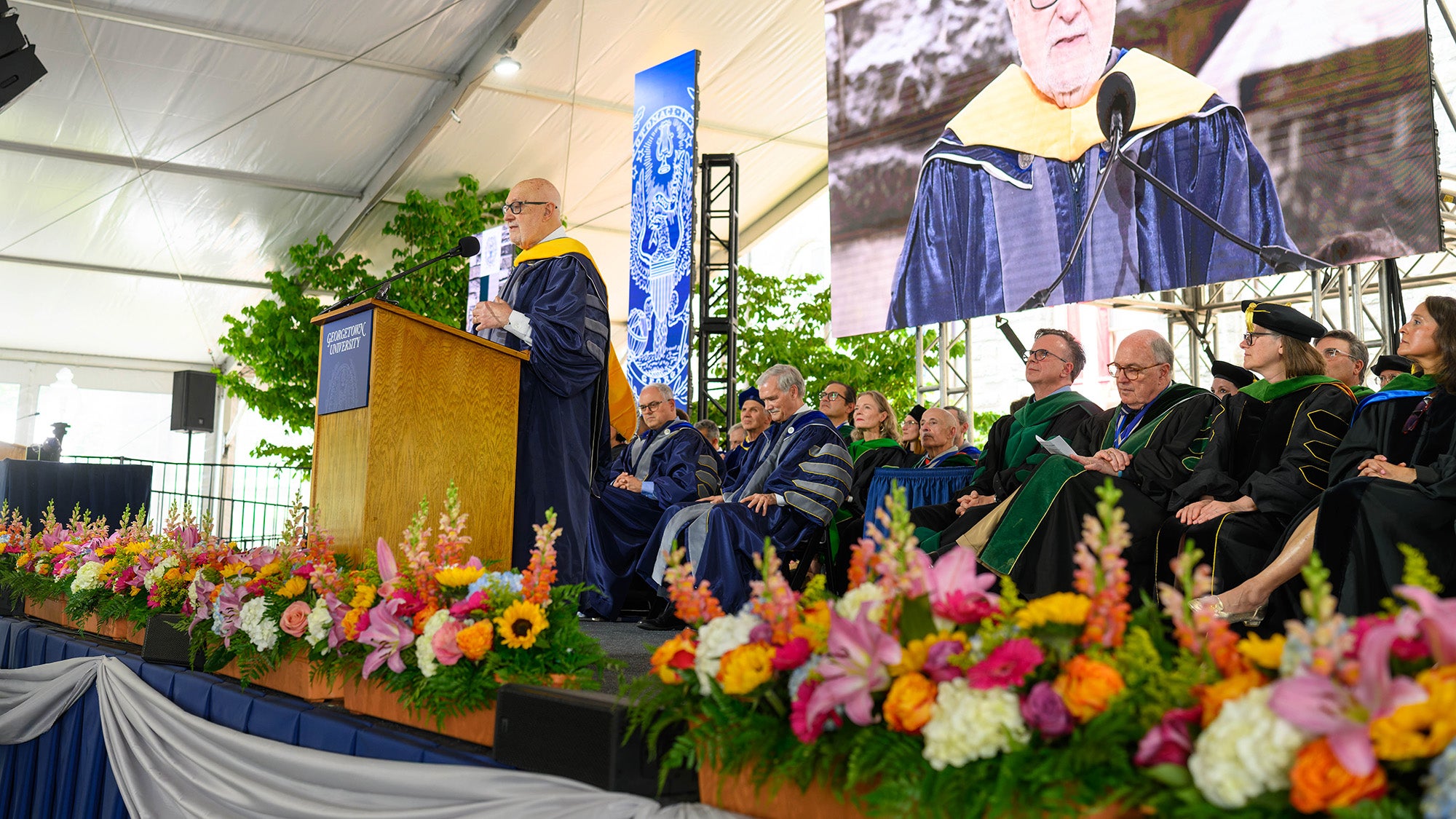 Pellegrino speaks from a podium onstage with a video monitor projecting his image behind him