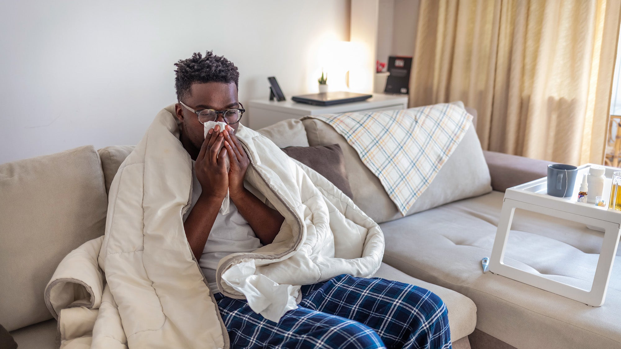 A Black man seated on a couch wrapped in a blanket blows his nose