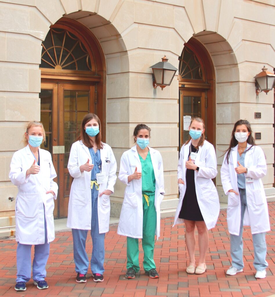 Five doctors in white coats and surgical masks stand outside the Med-Dent building giving a thumbs-up